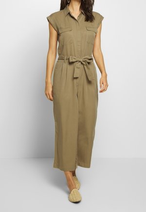 Kvinde iført ærmeløs olivengrøn buksedragt med bælte og vide ben samt beige slip-on sko, gående fremad.