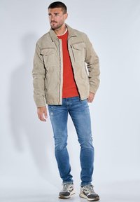 Beige Cordjacke aus Cord mit zwei Brusttaschen, getragen über einem roten, gerippten Pullover, kombiniert mit hellblauen Jeans und grauen Sneakern.