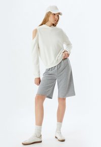 Sudadera blanca sin hombros, pantalones cortos grises, zapatillas blancas y calcetines beiges. Materiales suaves con un ajuste casual, que cuentan con capucha y aberturas laterales.