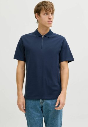Giovane uomo con polo blu navy a maniche corte con zip frontale, indossa jeans blu, guarda leggermente verso destra su sfondo semplice.
