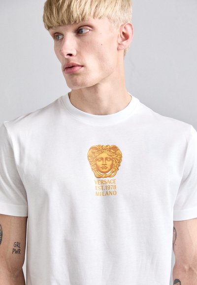 Blond man wearing a white T-shirt with a gold Versace Medusa logo and the text “VERSACE EST. 1978 MILANO” on the chest.
