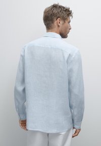 Chemise en lin bleu clair à manches longues, avec un empiècement au dos, des poignets à bouton et une texture lisse, tombant juste sous la taille.