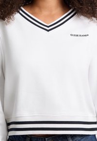 Bijela dukserica s V-izrezom i crnim prugastim rebrastim ovratnikom. Sadrži crne pruge na rubu i "GUESS JEANS" logo u crnom tekstu.
