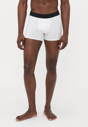 Mann, der barfuß steht und weiße Boxershorts mit einem schwarzen Bund trägt, vor einem schlichten hellen Hintergrund.