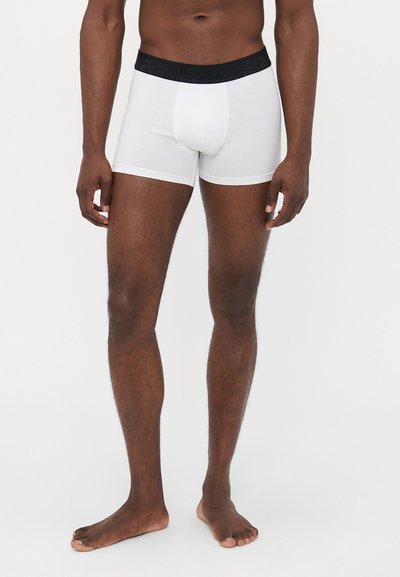 Homme debout pieds nus portant un boxer blanc à taille noire devant un fond léger uni.
