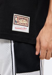 Μαύρη φανέλα μπάσκετ με ένα μπάλωμα "Hardwood Classics Mitchell & Ness Philadelphia PA" πάνω από λευκά και μαύρα διχτυωτά σορτς και ένα ορατό χέρι.