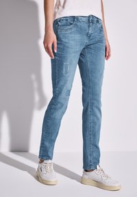Jean en denim bleu clair avec une coupe ajustée, présentant des écussons déchirés et un design classique à cinq poches. Porté avec des baskets blanches.
