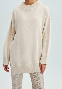 Krémový oversized svetr s ribbovaným výstřihem a manžetami. Pletená textura s volným střihem, doplněná třpytivými flitrovými kalhotami.