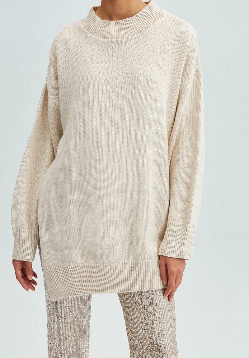Krémový oversized svetr s ribbovaným výstřihem a manžetami. Pletená textura s volným střihem, doplněná třpytivými flitrovými kalhotami.