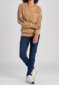 Tan sweatshirt med blå, röda och vita sidostreck, rund halsringning, ribbade ärmslut, kombinerad med mörkblå jeans och vita sneakers.