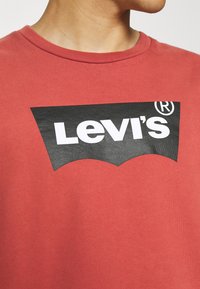 T-shirt in cotone rosso con logo nero Levi's caratterizzato da un bordo grafico curvo. Texture liscia e scollatura a girocollo.