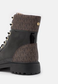 Bottine en cuir verni noir et motif marron avec œillets dorés, lacets sur le devant et semelle épaisse à crampons.