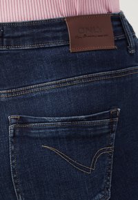 Mörkblå denimjeans med en brun läderetikett sydd i midjan och en distinkt sömnadsdesign på fickorna.
