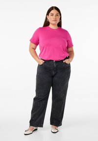 Fuchsia Kurzarm-T-Shirt kombiniert mit hochgeschnittenen dunklen Jeans. Schwarz-weiße spitze Schuhe. Model steht mit den Händen in den Taschen.