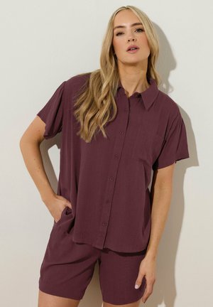 Blonde vrouw die een loszittend, kort mouwig overhemdbloesje en bijpassende korte broek in donkerpaars draagt, staand tegen een effen lichte muur.