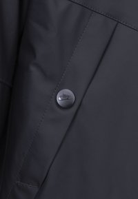 Nike gombík na tmavo námorníckej bunde, s hladkou textúrou, čistými líniami a kontrastným prešívaním v blízkosti detailu gombíka.