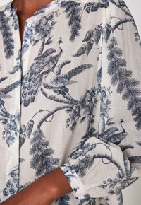 Donna che indossa una blusa bianca con dettagli in blu di un pavone e una stampa floreale, con bottoni sulla parte anteriore e maniche arrotolate.