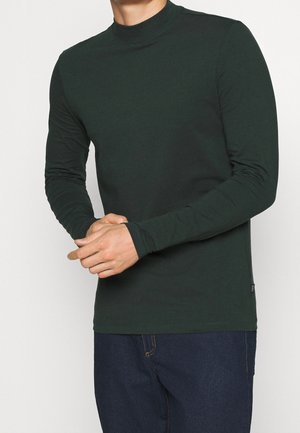 Uomo che indossa una maglia a maniche lunghe verde scuro con collo alla coreana e jeans blu scuro, con le mani intrecciate davanti all'altezza della vita.