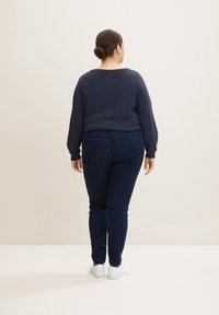 Sudadera azul marino con cuello redondo y mangas largas combinada con jeans ajustados de un azul oscuro. El modelo lleva zapatillas blancas y tiene un peinado recogido en un moño.