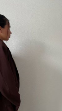 Oseba nosi dolg, temno rjav plašč in stoji ob teksturirani beli steni, z blago svetlobo in sencami.