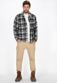 Ruudullinen flanellipaita musta-valkoisena, valkoisen T-paidan päällä. Beiget cargo-housut sivutaskuilla, yhdistettynä ruskeisiin nauhaliukuisiin kenkiin.