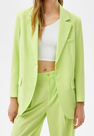 Frau trägt einen limettengrünen Blazer und passende Hose über einem weißen, bauchfreien Top, steht mit entspannten Händen an den Seiten.