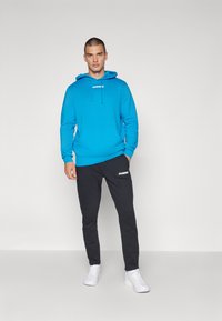 Felpa blu brillante con coulisse e tasca frontale, abbinata a pantaloni jogging neri con logo, completata da sneakers bianche.
