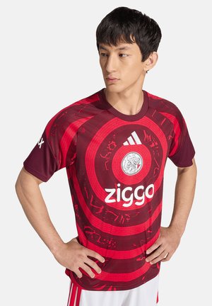 Hombre con cabello corto y negro, vistiendo una camiseta deportiva roja y granate con el logo de Ziggo y patrón circular, con las manos en las caderas, mirando hacia un lado.