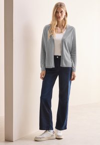 Grauer Cardigan über einem weißen Oberteil, kombiniert mit dunkelblauen, weiten Jeans und weißen Turnschuhen. Glatte Textur, lässiges Design.
