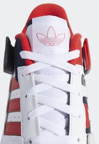 Gros plan d'une basket blanche, rouge et noire avec des lacets blancs et le logo trefoil rouge d’Adidas sur la languette.