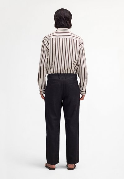 Osoba stojící zády k fotoaparátu, oblečená v dlouhém rukávu s vertikálními pruhy a v černých kalhotách, na bílém jednobarevném pozadí.