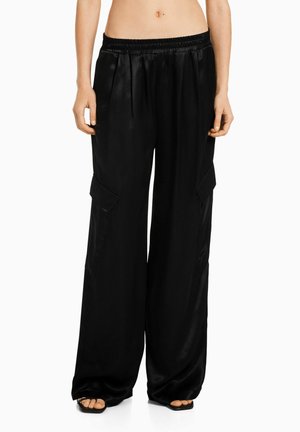 Pantalones anchos de satén negros con cintura elástica y bolsillos laterales con solapa, combinados con sandalias negras de punta abierta, mostrados desde la cintura hasta los pies.