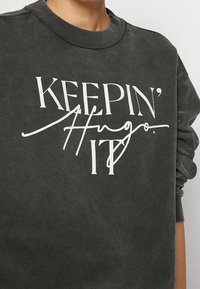 Felpa grigia scura con scollo rotondo. Presenta testo bianco: "KEEPIN' IT Hugo." Tessuto morbido, vestibilità casual e maniche leggermente oversize.