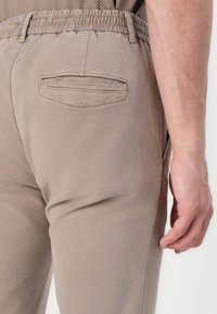 Stretchy beige pants with an elastic waistband and a small, back pocket featuring a double-stitched trim. Smooth fabric texture.
