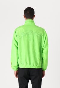 Casaco leve verde néon com punhos e barra elásticos, apresentando o texto em relevo "LACOSTE" nas costas e um design de gola alta.