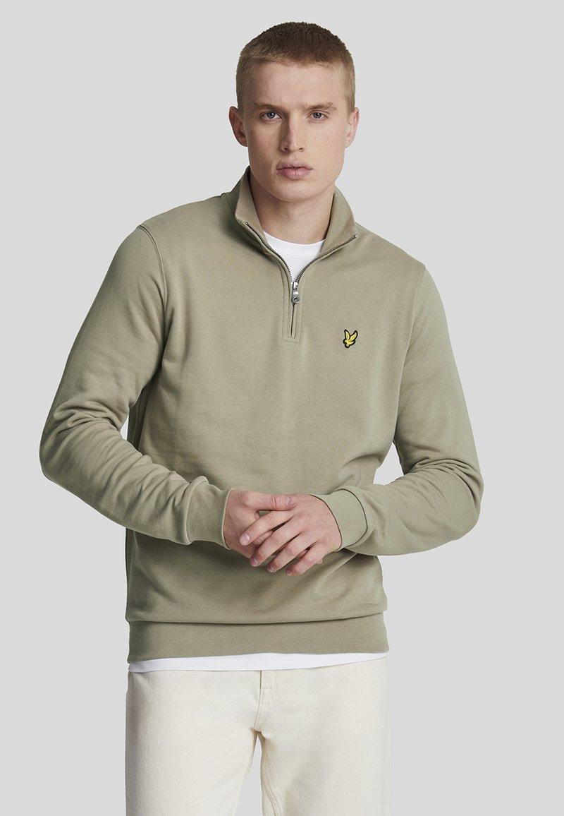 Pull en coton doux de couleur vert olive clair avec fermeture éclair sur le col, orné d'un logo jaune subtil sur la poitrine et de poignets et d'un ourlet côtelés.