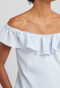 Lichtblauwe off-the-shoulder top met een gerimpelde halslijn, verticale strepen, zachte textuur en knoopaccenten op de mouwen.