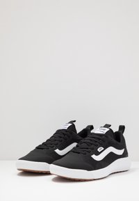 Ein Paar schwarze Vans Ultrarange Sneakers mit einem weißen Wellenstreifen, weißen Sohlen, schwarzen Schnürsenkeln und braunen Gummisohlen auf weißem Untergrund.