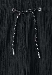 Un tissu noir texturé présente une taille à cordon avec une corde tressée noir et blanc contrastante et des embouts en métal.