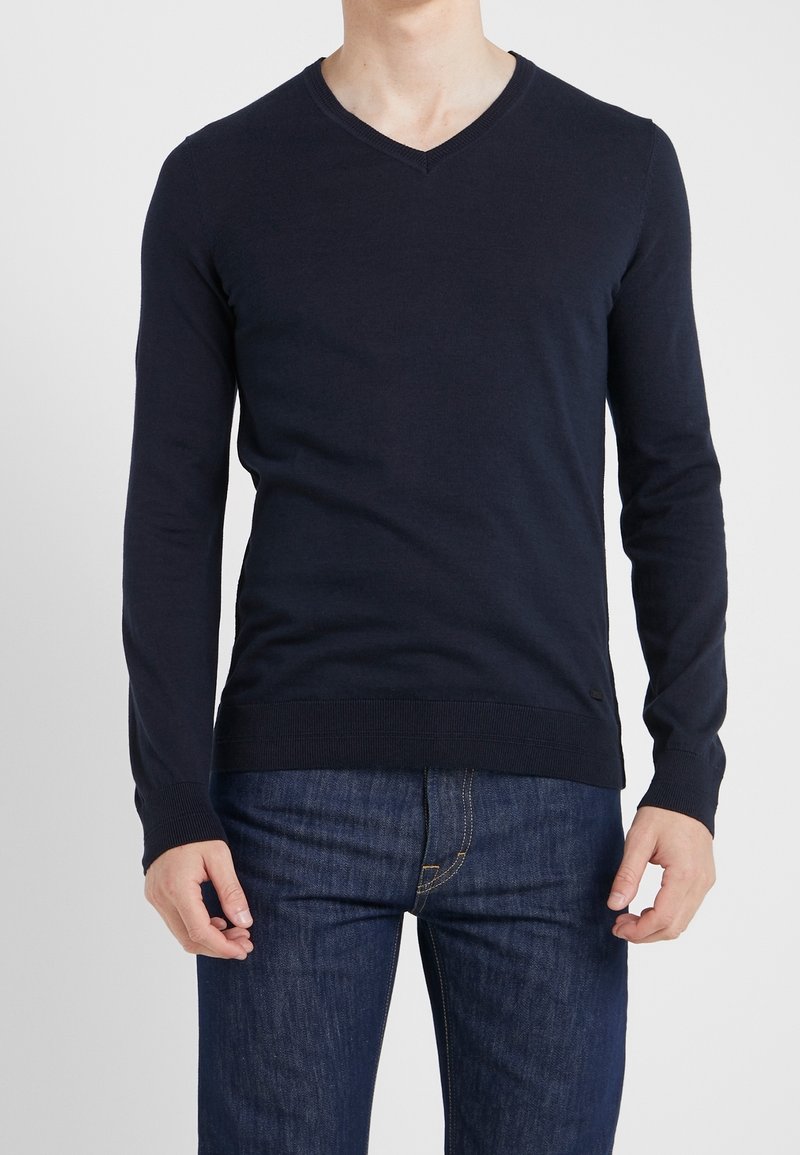 Marinblå, långärmad V-halströja i ett mjukt stickat tyg. Har ribbade ärmslut och nederkant, och är kombinerad med mörka denimjeans.