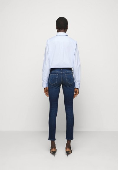 Jeans skinny azul escuro com cintura média, bolsos destacados e uma textura de denim suave, combinados com uma camisa azul clara.