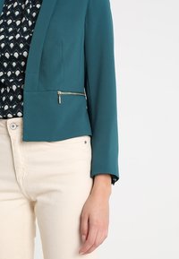 Žena v tyrkysovém zkráceném blejzru se zlatým zipem, pod nímž má vzorovanou černou blůzu, doplněná krémovými kalhotami s vysokým pasem, s uvolněnou rukou podél těla.