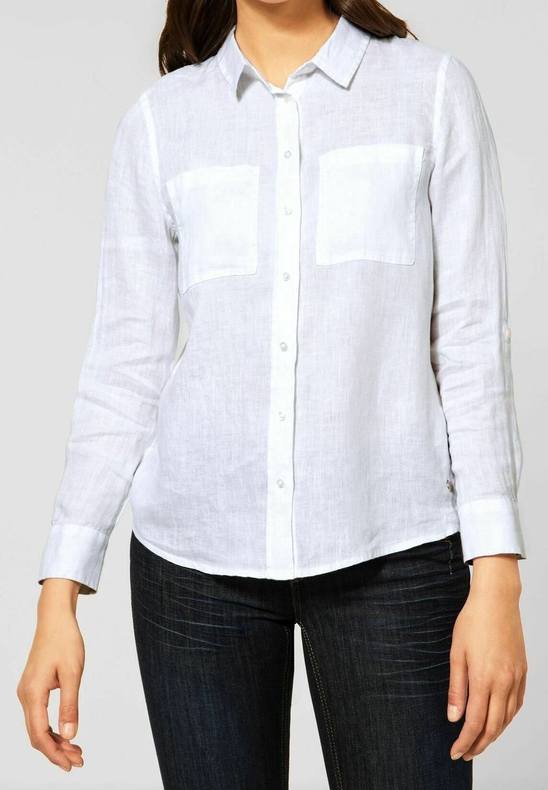 White linen shirt with long sleeves, button-down front, two chest pockets, and a curved hem, worn with dark blue jeans.