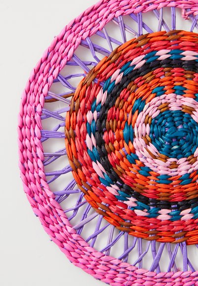 Circular woven mat featuring vibrant colors: pink, purple, orange, teal, and black. Made from natural fibers with a textured, layered design.