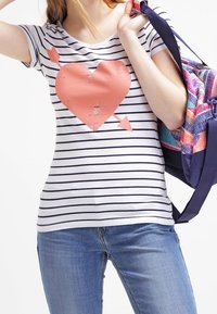 Camiseta a rayas blanca y azul marino con un gráfico de corazón coral y flechas, combinada con jeans de mezclilla y un bolso de patrón multicolor.