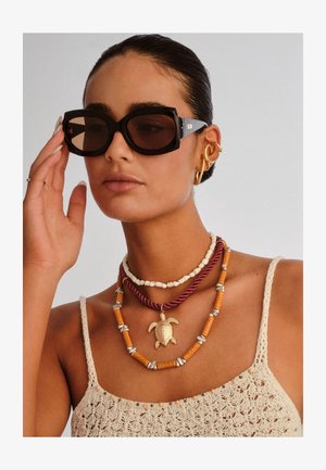 Ženska s temnimi sončnimi očali, plastenimi ogrlicami, vključno z eno z zlatim obeskom v obliki želve, zlatimi ušesnimi manšetami in kremno obrobljeno majico iz kvačkanja.