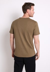 Man i en enkel brun kortärmad t-shirt och svarta byxor, vänd bort från kameran mot en ljus, enfärgad bakgrund.