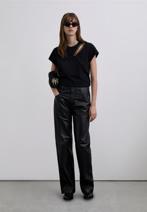 Camiseta negra de manga corta con detalle de recorte, combinada con pantalones de cuero negros de tiro alto. Modelo con gafas de sol negras, sosteniendo un bolso de mano.