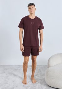 Viininpunainen puuvillainen t-paita pyöreällä kaula-aukolla ja pienellä logolla, yhdistetty vastaaviin shortseihin. Pehmeä koostumus, rento istuvuus, paljain jaloin.