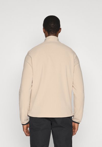 Person mit kurzen dunklen Haaren, der eine beige langärmlige Jacke und schwarze Hose trägt und vor einem schlichten weißen Hintergrund steht.